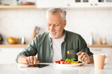 Cheerful Senior Man With Digital Tablet Reading News During Breakfast In Kitchen
