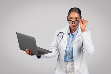 Glad shocked pretty millennial black lady doctor in white coat with laptop, with open mouth, take...