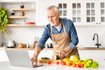 Smiling Elderly Man Looking Recipe On Laptop While Cooking Food In Kitchen