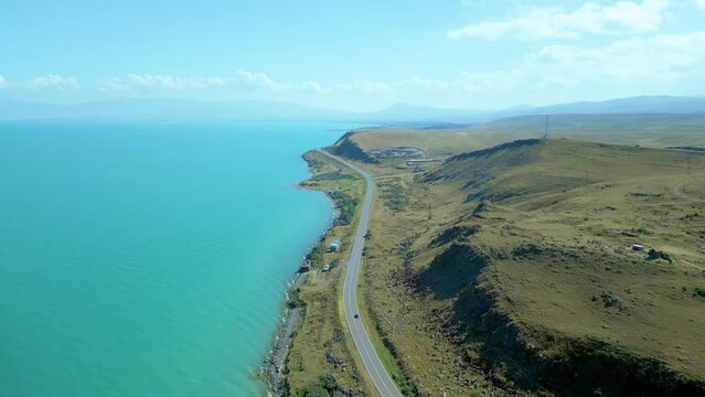 An aerial view of a large blue azure sea lake in clear sunny weather. On the shore of stony hills, in the distance high mountains in the clouds. Drone video, flying in an arc. Lake Sevan, Armenia.