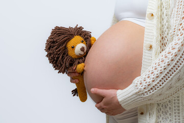 Close-up of a pregnant mother in white cloths holding a toy lion and placing her hand on her bare,...