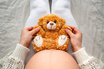 Close-up high angle shot of a cute toy bear sitting on mothers naked, round belly. Last month of...