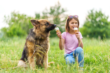 Smiling little caucasian girl eating ice cream cone and feeding it to her shepherd dog sitting on the grass in the park in summer. High quality photo, blurred background.