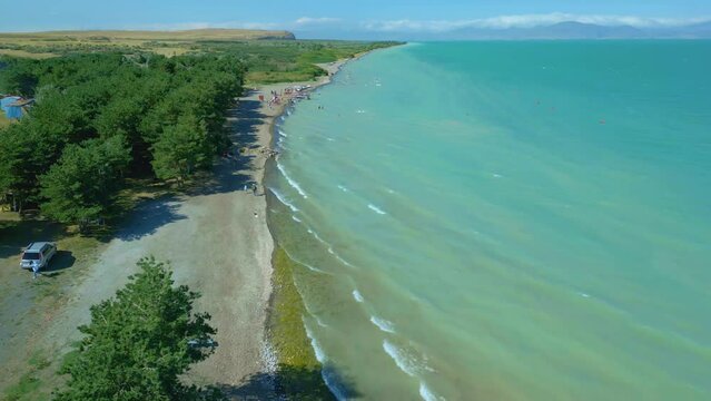 Aerial view of a large blue azure sea lake in clear sunny weather. People are swimming on the beach and pine trees are growing, boats are standing. Drone video, direct overflight. Lake Sevan, Armenia.