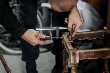 An adult male professional woodworker, a carpenter glues a chair, repairs, restores furniture indoors, workshop, clamping wooden products with a clamp. Close-up photography, work concept, portrait. - obrazy, fototapety, plakaty
