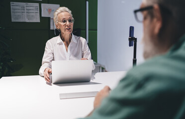 Senior Caucasian woman in business attire speaking in an office meeting, with a laptop in front of her and a senior male colleague listening, depicting a professional discussion