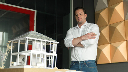 Portrait of smiling confident architect engineer looking at camera while standing with arm folded with house model placed on table. Professional businessman crossing arm. Design concept. Manipulator.
