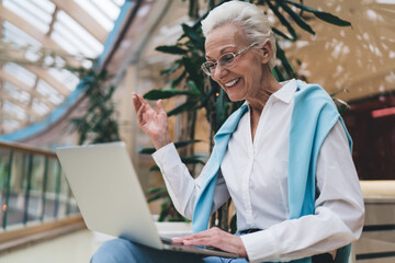 Elderly Caucasian woman in her 70s, smiling joyfully using laptop. She wear glasses, white shirt, ,...