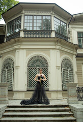 a 30-year-old girl in a sexy, designer, black dress, posing with a calla flower, against the background of a house. Photo shoot, stylish look in a translucent dress. Fashion trends, trends