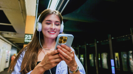 Young stunning woman using mobile phone and listening music playlist with wireless headphones while waiting on tram station.