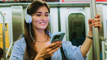 Female chatting online in mobile phone messenger watch video in metro. Young girl listening music in headphones and hold smartphone in subway car travel underground use wireless internet connection. - obrazy, fototapety, plakaty