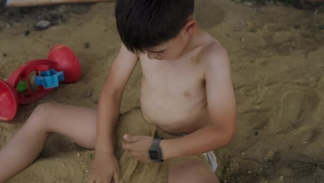 Boy sitting in only underpants on the sand in the garden of the house, covering his legs while digging with his hands in the sand. Summer vacation. Concept for