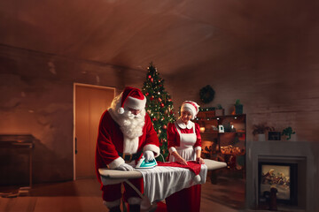 Préparatifs de noël : Père Noël repasse du linge sous la surveillance de Mère Noël dans leur salon devant la cheminée avec une décoration de noël et un sapin