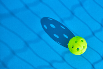 green pickleball ball on blue surface with the shadow of a net in front