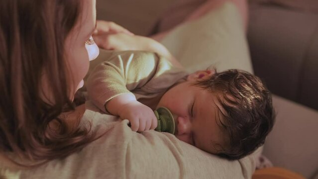 Mother smiles as holds baby in arms. Infant sucks pacifier and falls asleep in gentle embrace of mother under sunlight touching parent face
