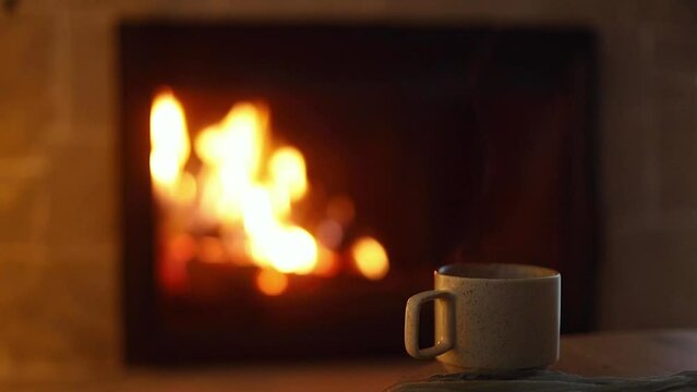 Stylish cup of warm tea on wooden table against cozy burning fireplace. Atmospheric eve at fireplace. Winter hygge. Autumn cozy evening footage