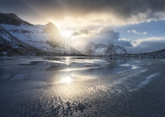 Beautiful snowy mountains and colorful sky with clouds and golden sunlight at sunset in winter in Lofoten islands, Norway. Landscape with rocks in snow, frozen sea coast, reflection in water. Nature