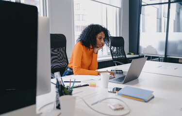 Deep in thought, an African American business consultant reviews complex data on laptop in coworking, orange sweater adding touch of warmth to professional atmosphere, moment of strategic planning.