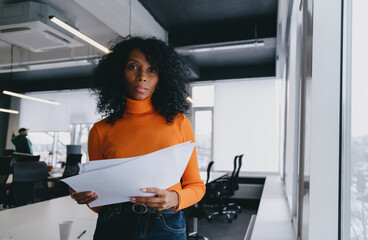 A perceptive analyst in a bold orange sweater scrutinizes financial reports in a bright office,...