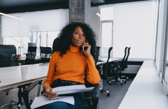 A Pensive Legal Advisor In A Vibrant Orange Sweater Evaluates Documents During A Phone Consultation In A Contemporary Office, Hinting At Discussions On Car Insurance Or Environmental Policies.