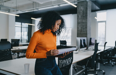 A Black woman in orange turtleneck sweater and blue jeans stands reading a report in modern office...