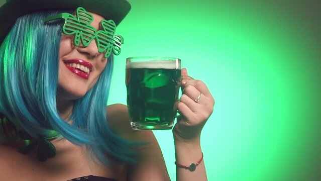 St. Patrick's Day leprechaun model girl in green hat holding mug of Green Beer pint and over green background, Dancing, Smiling. Patrick Day pub party, celebrating. Green beer. Ads. Slow motion