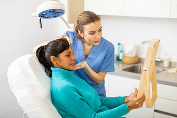 Positive Latin woman with mirror evaluating quality of cosmetological procedures. Her doctor, young European woman, standing beside.