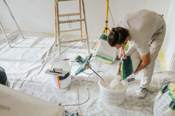 A builder is adding plaster in a bucket and making paste for plastering. - obrazy, fototapety, plakaty
