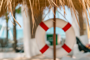 Intentionally blurred white red lifebuoy hang under tropical palm leaf umbrella, leafs are in focus