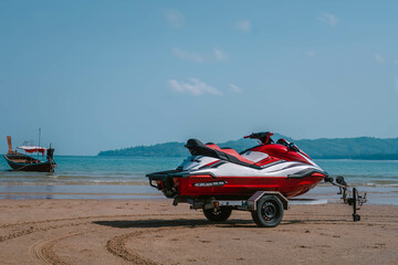 Red jet ski on trailer stands on sand beach, seashore and palm trees, morning time, tropical resort, Thailand. Rear side view
