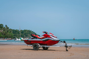 Red jet ski on trailer stands on sand beach, seashore and palm trees, morning time, tropical resort, Thailand. Side view