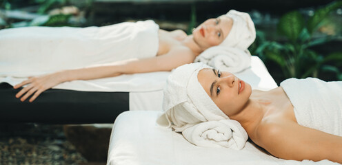 Obraz na płótnie Canvas Two beautiful young woman lie on spa bed with white towel while felling in deep relaxation rounded by relaxing and calming nature. Healthy and beauty concept. Blurring background. Tranquility.