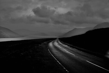 Greyscale of a road surrounded by a field to rocky smooth mountains