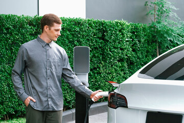 Young man travel with EV electric car charging in green sustainable city outdoor garden in summer...