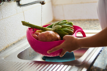 Close-up woman housewife washing fresh vegetables under running water, standing by stainless sink...