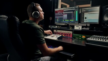 a focused male sound engineer wearing headphones sits at a mixing desk recording new track