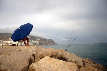 Man sitting under the umbrella and fishing from the shore against a rainbow in a cloudy weather