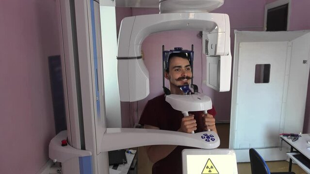 Smiling caucasian young adult man is sitting on computed tomography of head at dental clinic. Male person with curly mustache on x-ray diagnostic of teeth. Take care of health concept. Medicine theme.