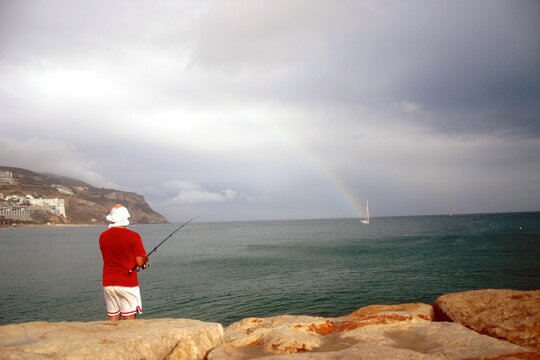 Man fishing from the shore against a rainbow in a cloudy weather