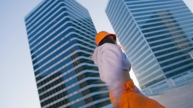 Hip-hop queen making twerk. Breakdancer woman enjoying sunset 4K. Joyful multi racial girl in sports outfit wearing trendy bright orange cargo pants and bucket hat dancing into music at city downtown