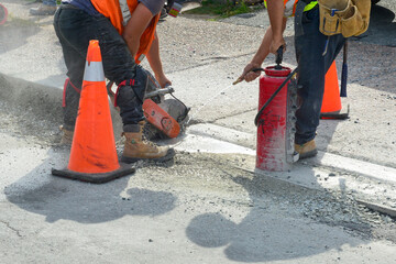 Two construction workers wearing belts cut a hole in a concrete sidewalk using a mechanical saw. An...