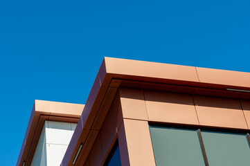 The roofs of a modern commercial building under blue sky and white clouds. The exterior of the new building is rusty, orange, and cream in color metal composite panels with black glass windows.  