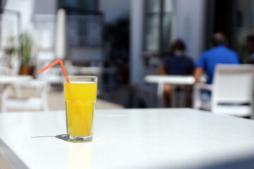 Glass of orange juice with plastic straw on white table in restaurant outdoor lounge zone in sunny weather