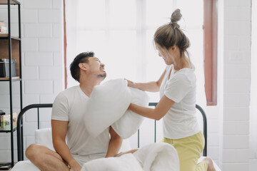 Funny couple angry wife hit husband with pillow in white bedroom.