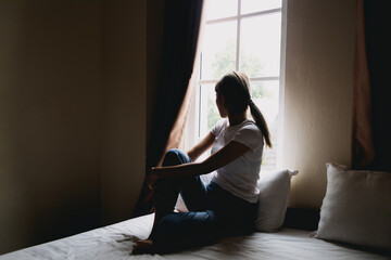 Lonely single asian woman sit alone in dark bedroom with light from curtain.