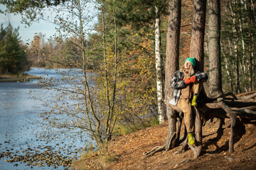 Carefree middle-aged woman sitting on pine tree root with eyes closed, turning face towards sun, sunbathing, warming, breathing fresh air, listening to birds, dreaming, enjoying warm autumn in nature