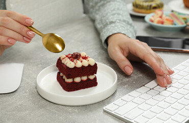 Bad habits. Woman eating piece of cake while working on computer at light grey table, closeup