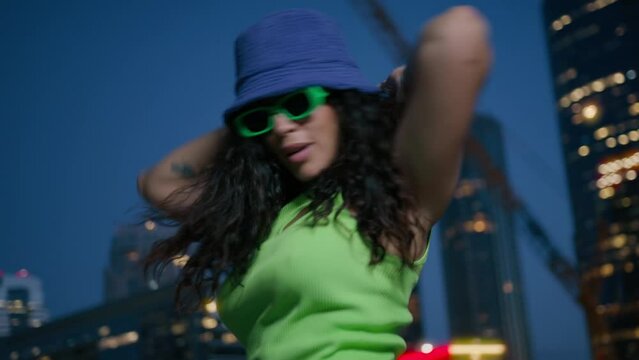 Cool sexy mixed race female modern dancer moves rhythmically 4K. Active african american black woman in neon green top and purple bucket hat dancing by urban lights background in downtown at night