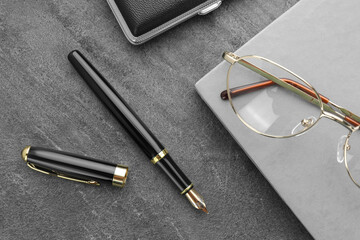 Stylish black fountain pen, glasses, notebook and cigarette case on grey textured table, flat lay