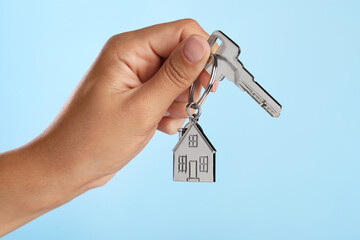 Woman holding key with metallic keychain in shape of house on light blue background, closeup